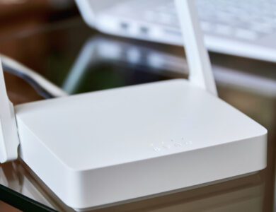 Weißer WLAN-Router auf einem Glastisch vor einem Laptop.
